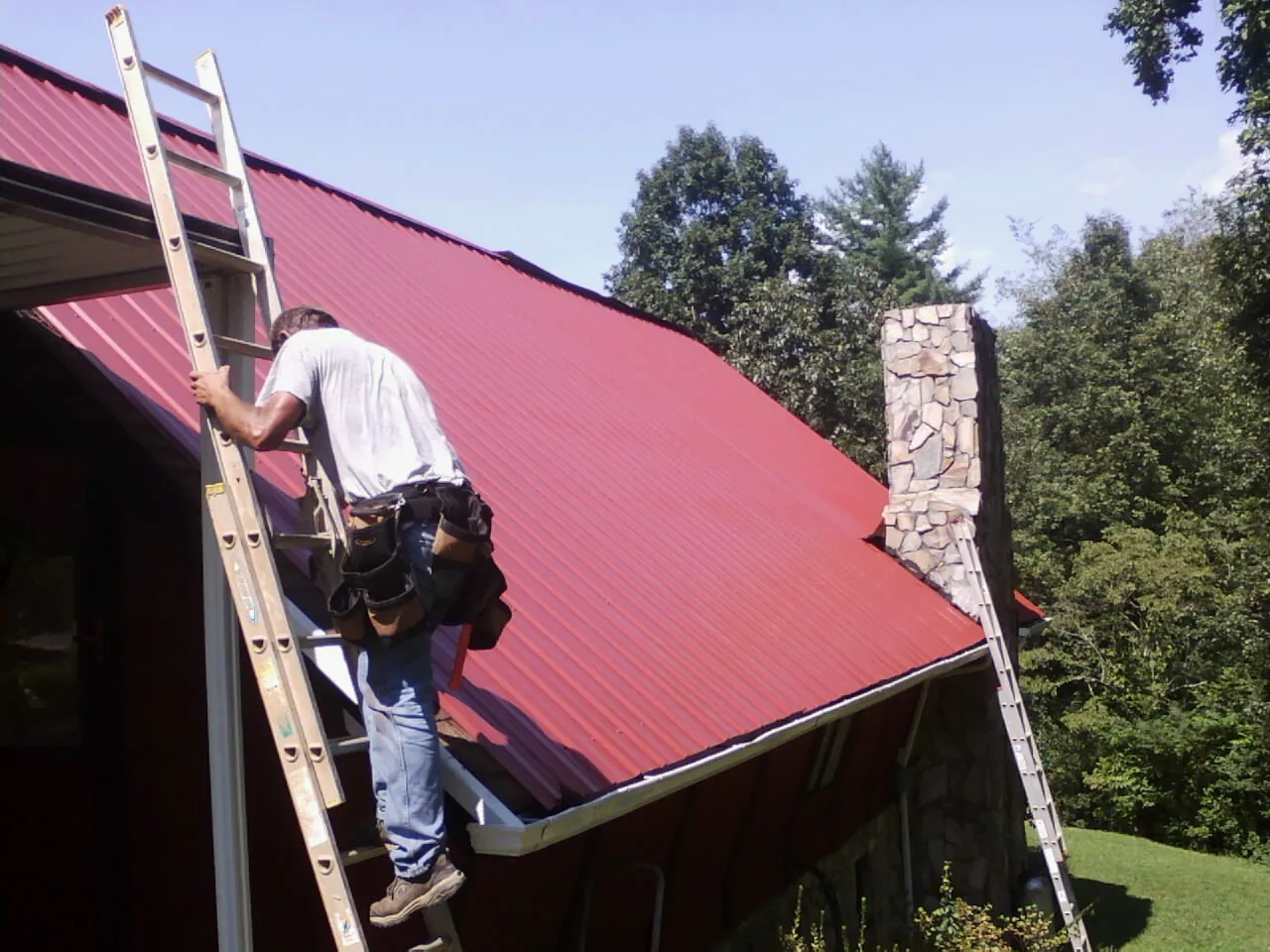 Roofer on a ladder beside a red metal roof and stone chimney — Soffit & Fascia Repair by Roofing Brothers in Bay Center